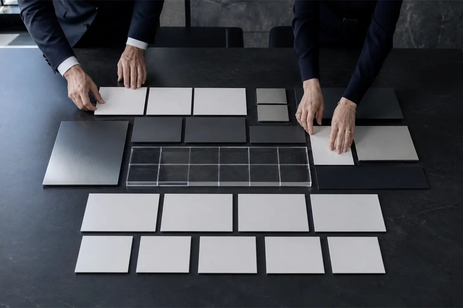 Two people arranging white and grey sample tiles on a black table
